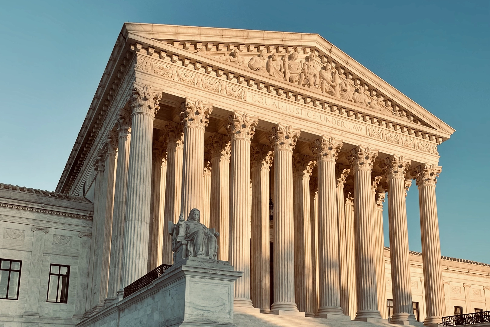 The image shows the facade of a neoclassical building with tall columns and a statue, under a clear blue sky.