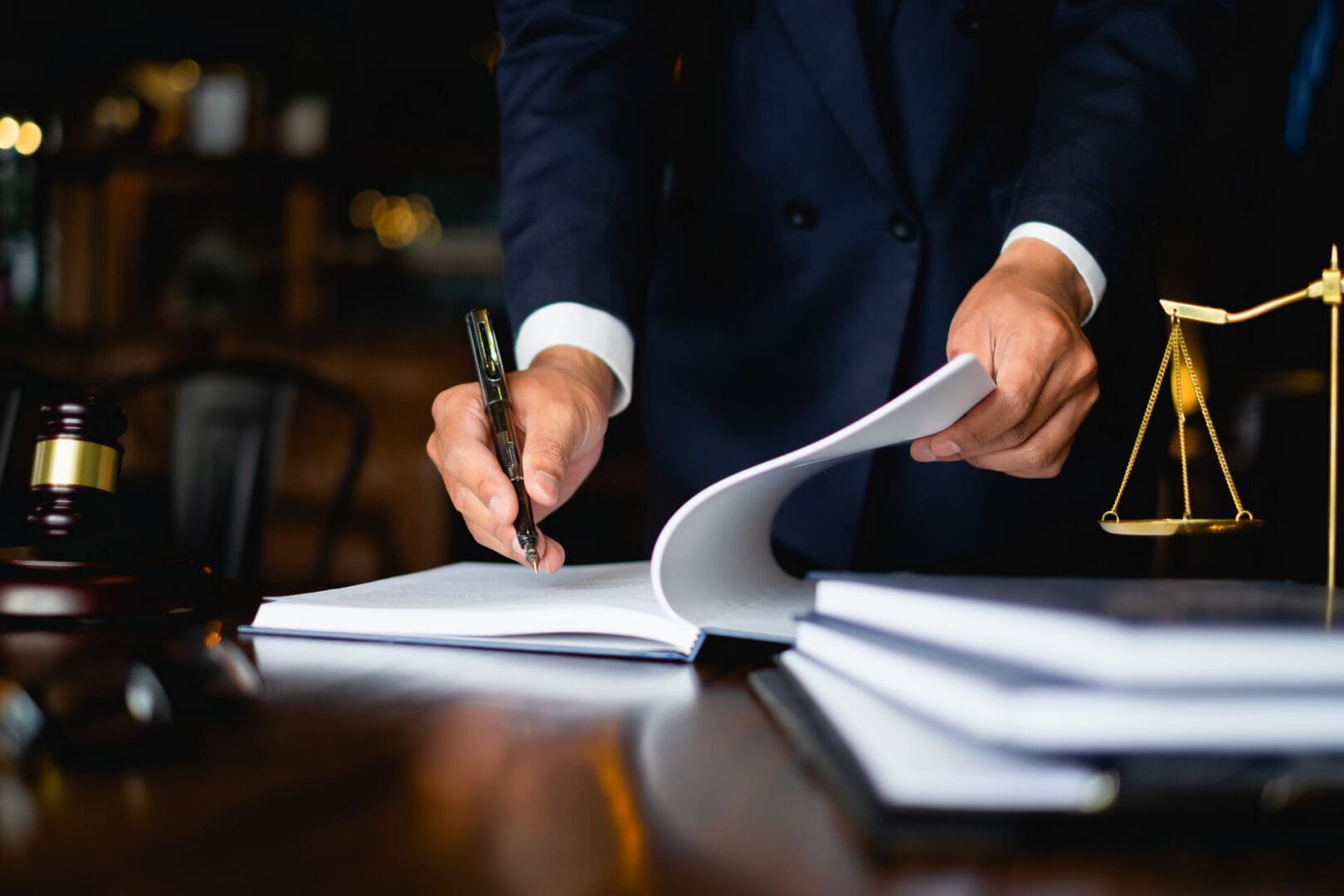 Person in a suit signing a document on a desk with legal books and a balance scale nearby.