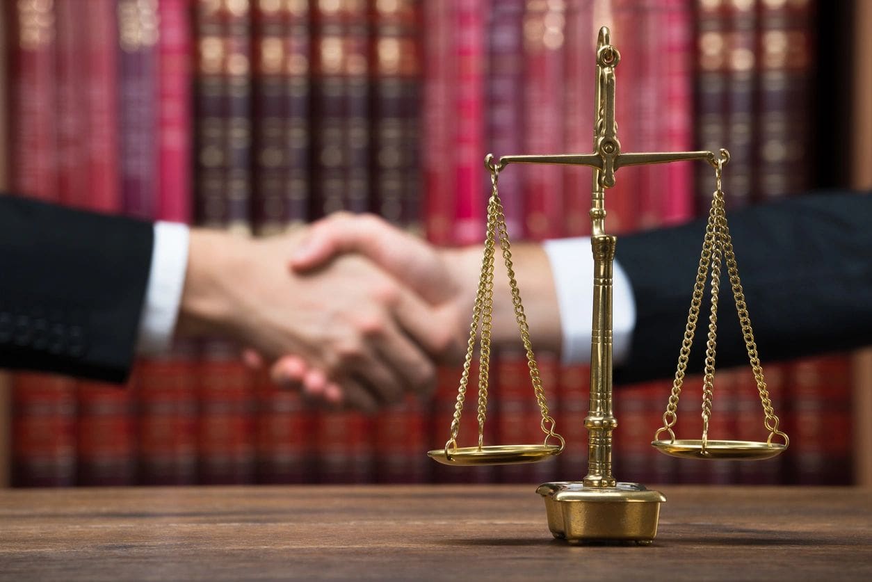 A pair of hands shaking behind a brass balance scale on a wooden table with shelves of books in the background.