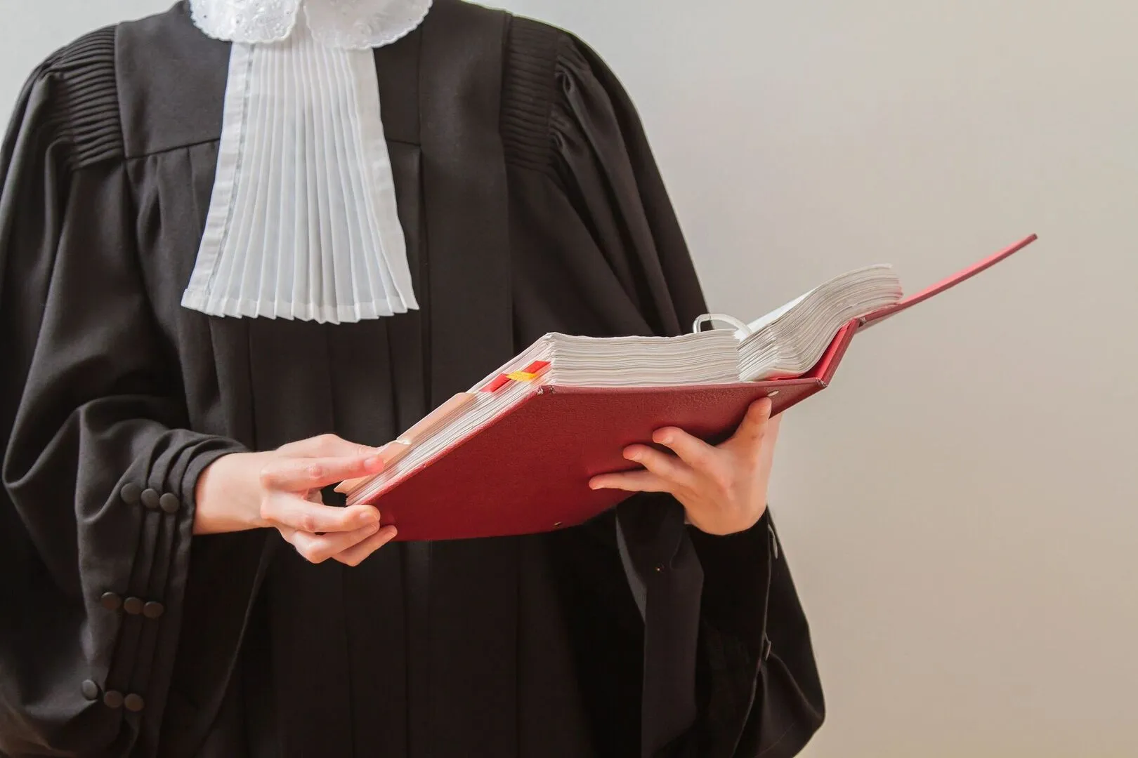 A person in a judge's robe holds and reads a large open book with a red cover.