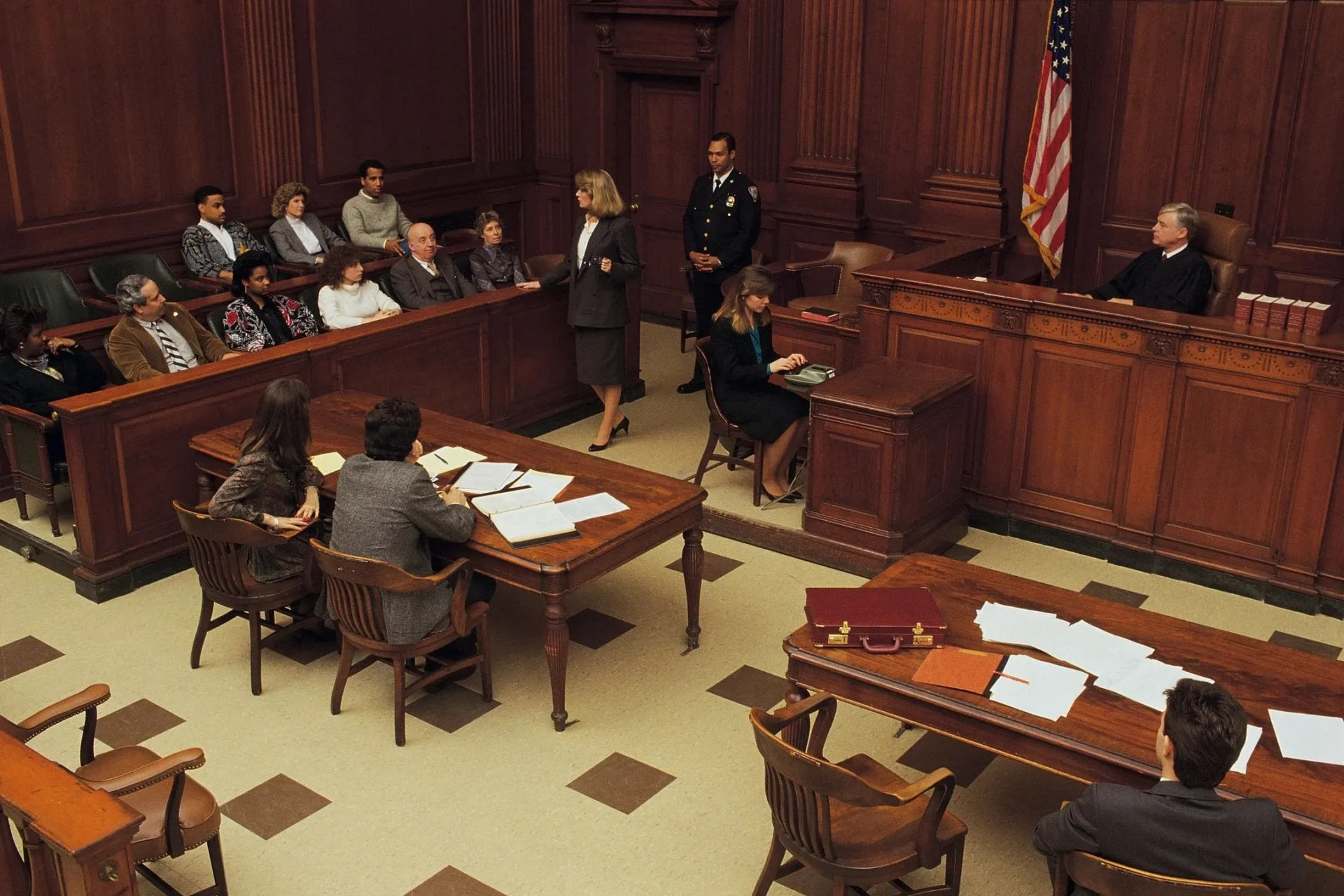 A courtroom scene with a lawyer addressing the jury, a judge seated at the bench, and papers on the tables.