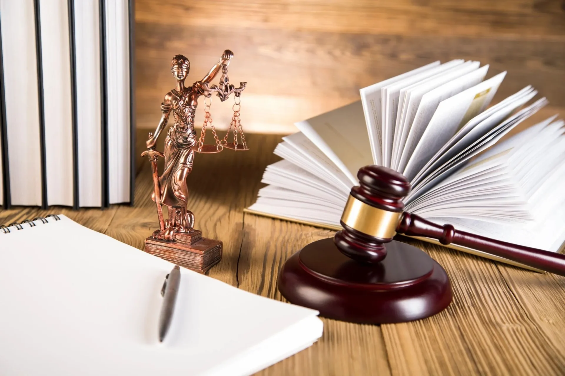 A gavel, Lady Justice statue, open book, closed books, and notebook with pen on a wooden desk.